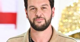 Claudio Caterisano smiles confidently, wearing a stylish beige jacket and layered necklaces, against a blurred background.