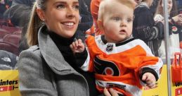 Cheerful woman holds baby wearing Flyers jersey, both enjoying a hockey game with ear protection for the little one.