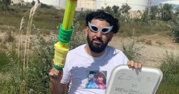 Steve Henni poses with a colorful water gun and a sign, surrounded by greenery and industrial background, exuding playful vibes.