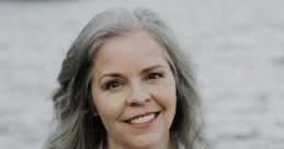 Smiling woman with gray hair, wearing a white shirt and turquoise necklace, by a serene water backdrop.