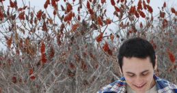 Gianni Matragrano enjoying winter outdoors, wearing a plaid shirt and surrounded by snow and vibrant red sumac.