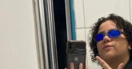 Person with curly hair wearing blue sunglasses, posing in a mirror, showcasing a stylish Jubs-themed outfit.