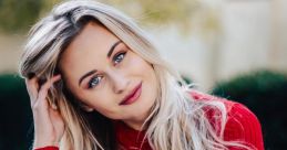 Smiling young woman with long blonde hair, wearing a red sweater, poses outdoors with a relaxed and inviting expression.