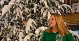 A woman in a green sweater and black pants holds a glass of wine, surrounded by snow-covered trees.