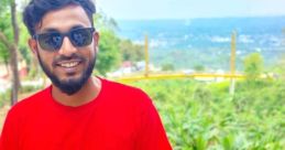 Smiling man in a red t-shirt and sunglasses, surrounded by lush greenery, enjoying a scenic outdoor view.