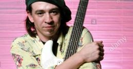 Musician embracing a white guitar, wearing a colorful shirt and a black beret against a soft pink background.