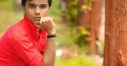 Akarsh Sharma in a red shirt and black pants, sitting pensively in a green park setting, showcasing a relaxed vibe.