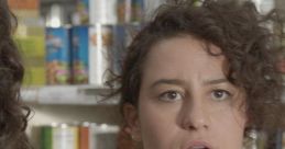 Ilana Glazer looks astonished while shopping in a grocery store, surrounded by colorful canned goods.