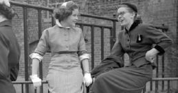 Two stylish women laughing in 1950s fashion, sitting on a fence with high heels nearby, capturing a joyful moment.