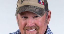 Larry the Cable Guy smiles and points energetically, wearing a plaid shirt and camo cap, embodying his comedic persona.