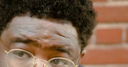 A thoughtful individual with curly hair and glasses, wearing a Dizzyeight-themed shirt, against a brick wall backdrop.