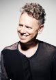 Martin L. Gore smiling, showcasing his iconic curly hairstyle, dressed in a stylish black shirt against a neutral background.