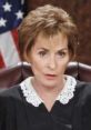 Judge Judy in a black robe with a lace collar, serious expression, sitting in court with an American flag backdrop.