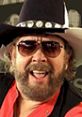 Hank Williams Jr. smiles in sunglasses and a cowboy hat, showcasing his iconic country music style and vibrant personality.