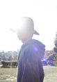 Akita Hiromu in silhouette, wearing a hat, with sunlight creating a dramatic effect outdoors. Amazarashi vibe captured.