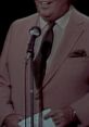 Edward Herlihy narrating a newsreel in 1965, wearing a suit and smiling, speaking into a microphone on stage.