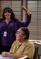 A cheerful woman holding a corndog playfully interacts with an elderly man at a table, capturing the humor of B Positive Season 2.