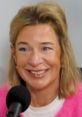 Katie Hopkins smiling while speaking into a microphone, wearing a pink sweater in a studio setting.