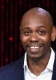 Dave Chappelle smiling broadly, showcasing his charismatic personality on stage, surrounded by vibrant background lights.