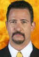 Jim Rome, sports talk show host, wearing a suit with a confident expression against a vibrant orange background.