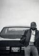 Kendrick Lamar poses confidently by a classic car, embodying urban style and creativity in a striking black-and-white setting.