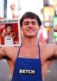 Shirtless man in Times Square holding a magazine, wearing an apron with 'BETCH', promoting QCP brand lifestyle.