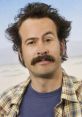 Character from "My Name is Earl" with a mustache and messy hair, wearing a plaid shirt and blue t-shirt, smiling warmly.