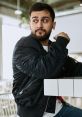 Stylish young man with a beard wearing a black bomber jacket, sitting at a modern cafe table, looking thoughtfully.