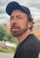 Benny Benassi with a beard, wearing a black cap and shirt, gazes thoughtfully outdoors against a cloudy sky backdrop.