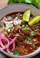 Savory beef stew topped with onions, cilantro, lime wedges, and pink pickled onions, perfect for a hearty meal.