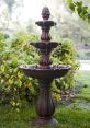 Elegant three-tiered fountain with water cascading over decorative bowls, surrounded by lush greenery and vibrant plants.