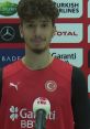 Berkcan smiles during a press conference, wearing a red sports jersey with Turkish flag, promoting basketball in Turkey.