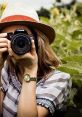 Young woman with a Canon camera in a green garden, capturing nature with a stylish hat and patterned blouse.