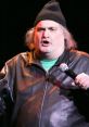 Artie Lange performs stand-up comedy on stage, wearing a black beanie and leather jacket, engaging the audience with humor.