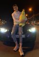 Young man in a hoodie poses with a plush banana on a car, showcasing casual nighttime style and urban vibes.