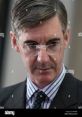 Jacob Rees-Mogg in a formal setting, wearing glasses, a striped shirt, and a tie, looking thoughtful and engaged.