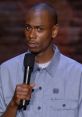 Dave Chappelle performing stand-up comedy, holding a microphone, with a focused expression, engaging the audience.