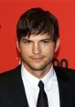 Ashton Kutcher at an event, showcasing his signature style with a sharp suit and confident expression against a vibrant backdrop.