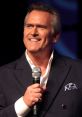 Bruce Campbell smiles while speaking into a microphone at a live event, showcasing his charismatic presence.