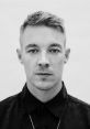 Portrait of a man with short hair and a serious expression, wearing a dark shirt and necklace, with a minimalistic backdrop.