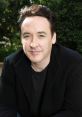 John Cusack smiles casually while wearing a black jacket, surrounded by greenery, showcasing a relaxed vibe.