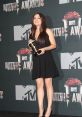 Actress on stage holding an award at the MTV Movie Awards, wearing a black dress with a confident smile.