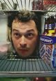 A man peering out from a refrigerator, surrounded by food and Bud Light bottles, reflecting humor and surprise.