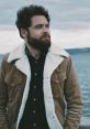 Sol Rosenberg outdoors, wearing a stylish brown jacket with a shearling collar, gazing thoughtfully at the scenic background.