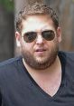 Jonah Hill wearing sunglasses and a black V-neck shirt, captured candidly outdoors, showcasing his unique style.