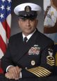 Portrait of a uniformed Navy officer with medals, against a backdrop featuring the American flag and an emblematic banner.