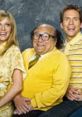 Cast of "It's Always Sunny in Philadelphia" wearing yellow outfits, smiling together in a playful pose against a gray backdrop.