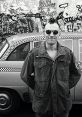 Travis Bickle stands in front of a classic taxi, embodying the iconic look from the movie "Taxi Driver" with urban graffiti backdrop.