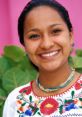 Smiling woman wearing traditional embroidered blouse and colorful jewelry, surrounded by lush greenery and pink background.