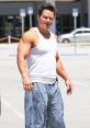 Mark Wahlberg in a white tank top and patterned pants, showcasing his muscular build in a casual outdoor setting.